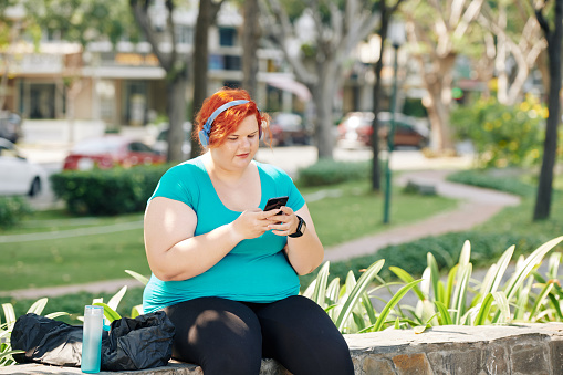 Overweight young woman with bright red hair resting in city park on sunny day and checking social media on her smartphone