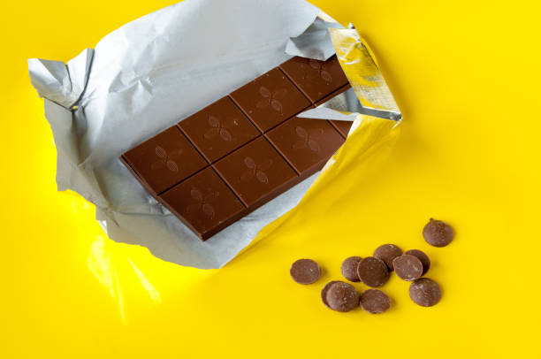 istock Milk chocolate bar in silver foil and chocolate drops on a yellow background 1212664069