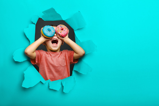 Happy cute boy is having fun played with donuts on black background wall. Bright photo of a child. Colored donuts