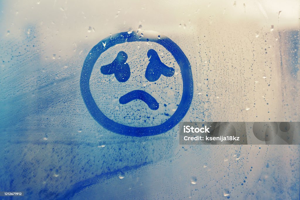 drawn sad face on foggy glass window raindrops blue color concept photo Hand drawn melancholy face painted on window flooded with raindrops on blue background Sadness Stock Photo