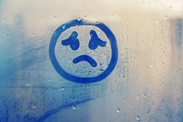 drawn sad face on foggy glass window raindrops blue color concept photo Hand drawn melancholy face painted on window flooded with raindrops on blue background anthropomorphic face photos stock pictures, royalty-free photos & images