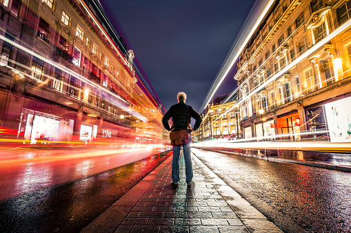 Wide angle color image depicting a rear view of one man standing in the middle of a busy street in central London illuminated at night. The man is surrounded by blurred motion of traffic light trails on both sides of the road. The man is on his own and there are no other people in this busy city street, due to the long exposure used. The man is in his 50s or 60s and is wearing casual clothing - blue denim jeans, a green jacket and a brown leather messenger bag.