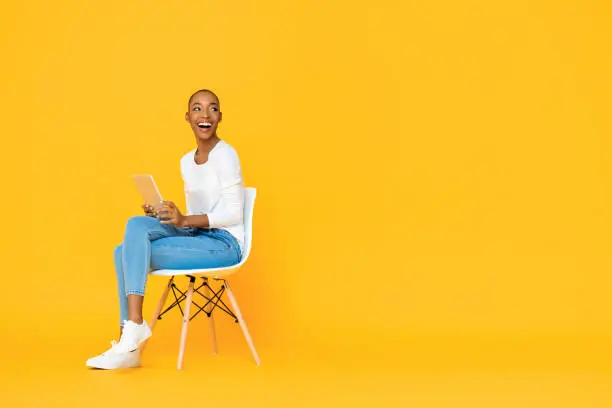 Photo of Trendy smiling African American woman sitting on a chair using tablet computer thinking and looking at empty space aside isolated yellow background
