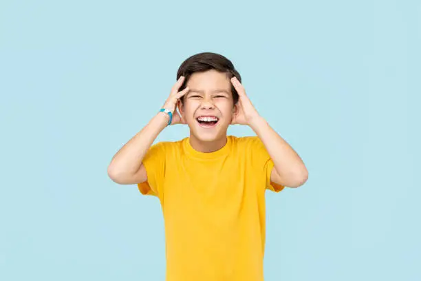 Happy smiling Asian boy with hands squeezing head isolated on light blue background