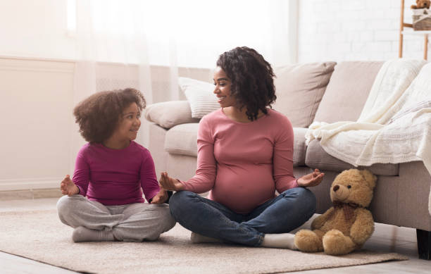 mère noire enceinte et sa petite fille pratiquant youga à la maison - floor women sitting yoga photos et images de collection