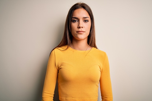 Young beautiful brunette woman wearing yellow casual t-shirt over white background with serious expression on face. Simple and natural looking at the camera.