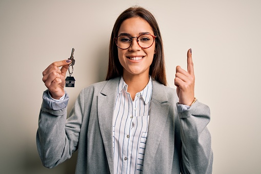 Young beautiful brunette house agent woman holding key home over white background surprised with an idea or question pointing finger with happy face, number one