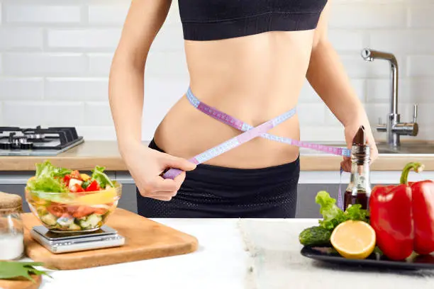 Photo of Young sports girl with naked torso measuring her waist with measuring tape in the kitchen. Diet and weigh loss concept