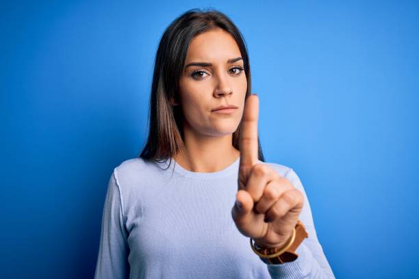 young beautiful brunette woman wearing casual sweater standing over blue background pointing with finger up and angry expression, showing no gesture - behavior women anger pointing imagens e fotografias de stock