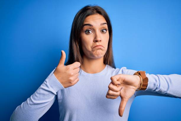 young beautiful brunette woman wearing casual sweater standing over blue background doing thumbs up and down, disagreement and agreement expression. crazy conflict - thumbs up human thumb human hand conflict imagens e fotografias de stock