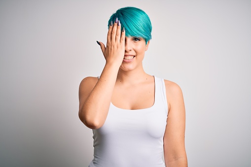 Young beautiful woman with blue fashion hair wearing casual t-shirt over white background covering one eye with hand, confident smile on face and surprise emotion.