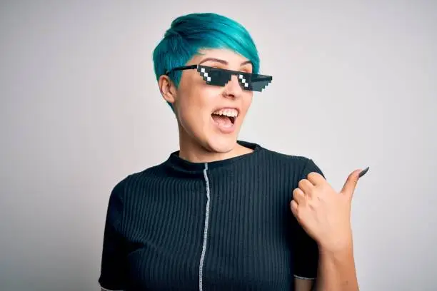 Young woman with blue fashion hair wearing thug life sunglasses over white background smiling with happy face looking and pointing to the side with thumb up.