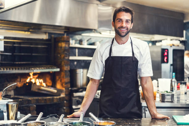Confident male chef in commercial kitchen Portrait of smiling mid adult chef in commercial kitchen. Confident male cook is standing at counter. He is working in restaurant. three quarter length photos stock pictures, royalty-free photos & images
