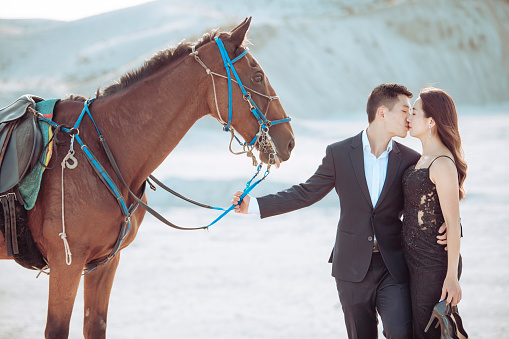 Young woman standing next to white Arabian horse resting in wooden stables box, kissing it eyes closed, sun shines on them, closeup detail