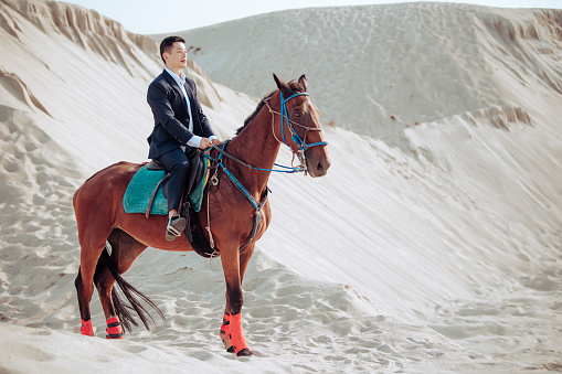 an asian chinese male with formal clothing riding a horse in the desert sand dune of melaka during sunset