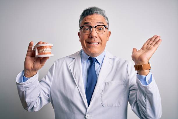 istock Middle age handsome grey-haired dentist man holding plastic teeth over white background very happy and excited, winner expression celebrating victory screaming with big smile and raised hands 1212691495