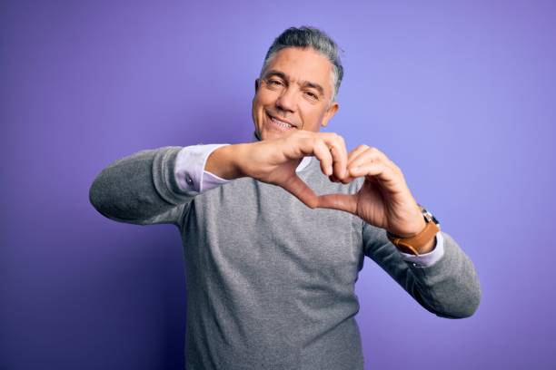 hombre guapo de la edad media con suéter elegante sobre fondo púrpura sonriendo en amor haciendo forma de símbolo de corazón con las manos. concepto romántico. - made man object fotografías e imágenes de stock