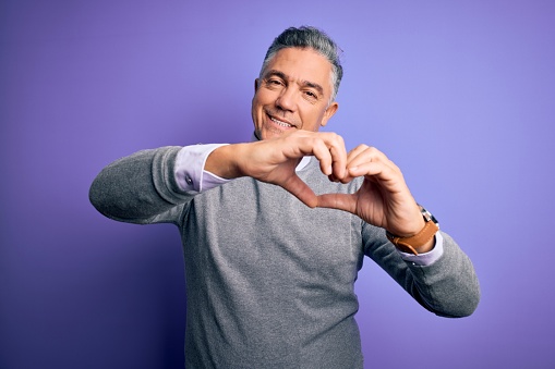 Middle age handsome grey-haired man wearing elegant sweater over purple background smiling in love doing heart symbol shape with hands. Romantic concept.