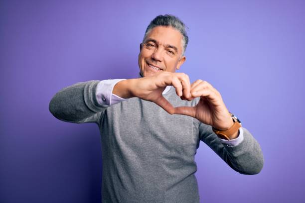 middle age handsome grey-haired man wearing elegant sweater over purple background smiling in love doing heart symbol shape with hands. romantic concept. - made man object imagens e fotografias de stock
