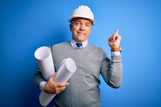 istock Middle age handsome grey-haired architect man wearing safety helmet holding blueprints cheerful with a smile on face pointing with hand and finger up to the side with happy and natural expression 1212692429