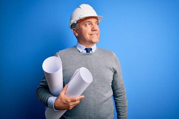 Middle age handsome grey-haired architect man wearing safety helmet holding blueprints looking away to side with smile on face, natural expression. Laughing confident.