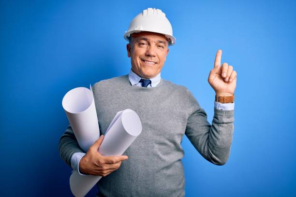 istock Middle age handsome grey-haired architect man wearing safety helmet holding blueprints with a big smile on face, pointing with hand finger to the side looking at the camera. 1212692481