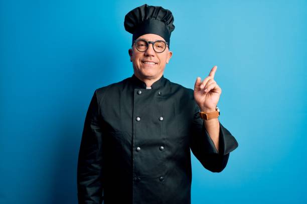 istock Middle age handsome grey-haired chef man wearing cooker uniform and hat with a big smile on face, pointing with hand finger to the side looking at the camera. 1212692575