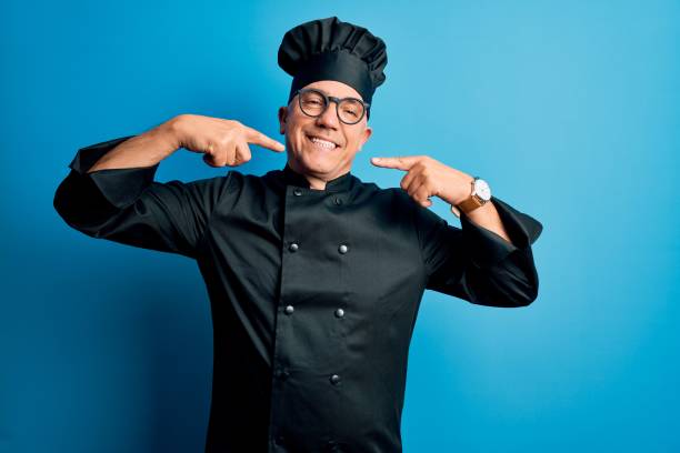 istock Middle age handsome grey-haired chef man wearing cooker uniform and hat smiling cheerful showing and pointing with fingers teeth and mouth. Dental health concept. 1212692586