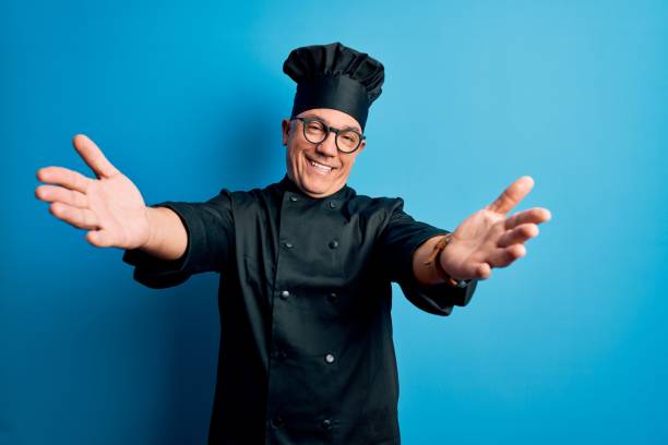 istock Middle age handsome grey-haired chef man wearing cooker uniform and hat looking at the camera smiling with open arms for hug. Cheerful expression embracing happiness. 1212692607