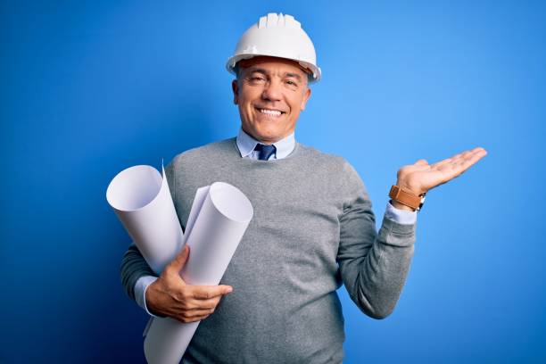 istock Middle age handsome grey-haired architect man wearing safety helmet holding blueprints smiling cheerful presenting and pointing with palm of hand looking at the camera. 1212692719