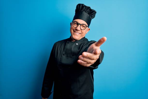 istock Middle age handsome grey-haired chef man wearing cooker uniform and hat smiling cheerful offering palm hand giving assistance and acceptance. 1212692761