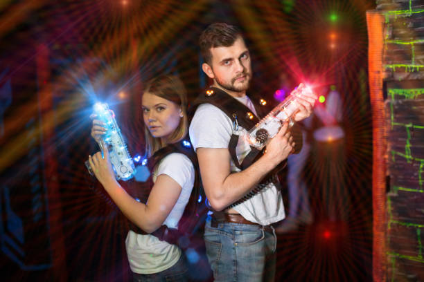 Couple standing back to back in colorful laser beams, holding guns during lasertag game in labyrinth