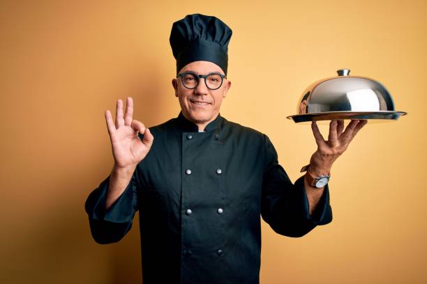 istock Middle age handsome grey-haired waiter man wearing cooker uniform and hat holding tray smiling positive doing ok sign with hand and fingers. Successful expression. 1212692977