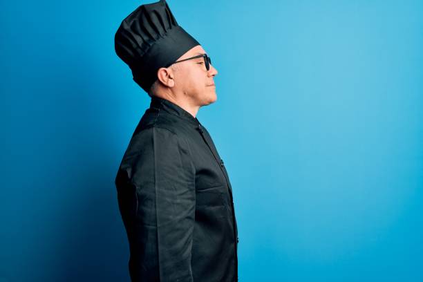 Middle age handsome grey-haired chef man wearing cooker uniform and hat looking to side, relax profile pose with natural face and confident smile.