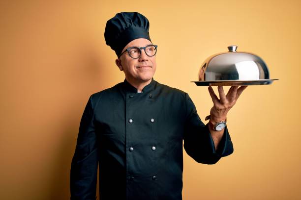istock Middle age handsome grey-haired waiter man wearing cooker uniform and hat holding tray smiling looking to the side and staring away thinking. 1212692987