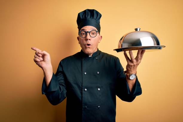 istock Middle age handsome grey-haired waiter man wearing cooker uniform and hat holding tray Surprised pointing with finger to the side, open mouth amazed expression. 1212693021