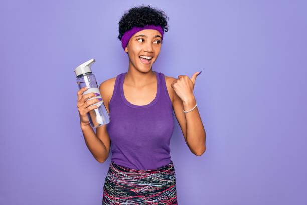 Young beautiful african american sporty woman wearing sportswear drinking bottle of water pointing and showing with thumb up to the side with happy face smiling Young beautiful african american sporty woman wearing sportswear drinking bottle of water pointing and showing with thumb up to the side with happy face smiling sportsman professional sport side view horizontal stock pictures, royalty-free photos & images