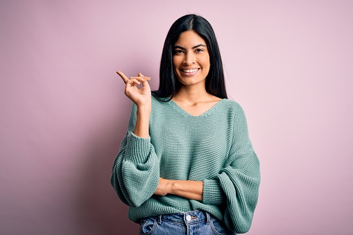 Young beautiful hispanic fashion woman wearing green sweater over pink background with a big smile on face, pointing with hand and finger to the side looking at the camera.