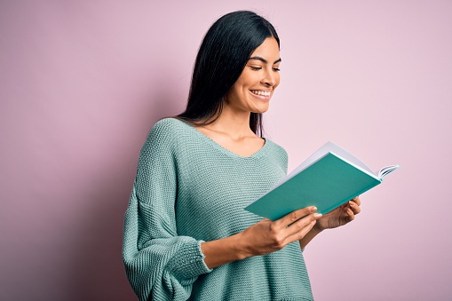 Young beautiful hispanic student woman reading a book over pink isolated background with a happy face standing and smiling with a confident smile showing teeth