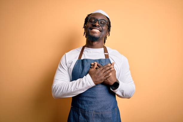 jeune homme d’autocuise afro-américain utilisant le tablier et au-dessus du fond jaune isolé souriant avec des mains sur la poitrine avec les yeux fermés et le geste reconnaissant sur le visage. concept de santé. - chef restaurant cooking african descent photos et images de collection