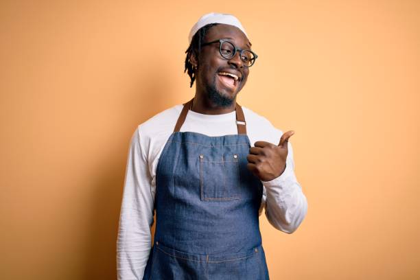 エプロンを身に着け、孤立した黄色の背景を身に着けている若いアフリカ系アメリカ人の調理器具の男は、幸せな顔を見て、親指を上げて横を指しています。 - baker occupation ストックフォトと画像
