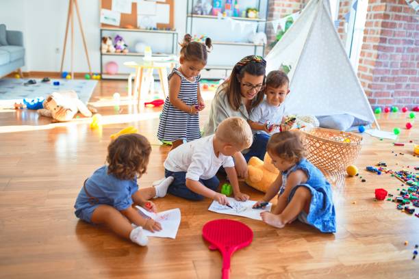 hermoso maestro y grupo de niños pequeños sentados en el suelo dibujando usando papel y lápiz alrededor de un montón de juguetes en el jardín de infantes - preschooler fotografías e imágenes de stock