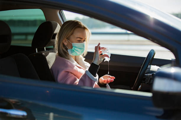 donna in maschera protettiva seduta in auto su strada, usando disinfettante per le mani. - driving women rear view sitting foto e immagini stock