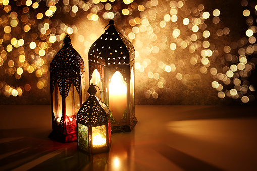 Ornamental Arabic lanterns with glittering bokeh lights. Burning candles on table glowing at night. Festive greeting card, invitation for Muslim holiday Ramadan Kareem,golden Iftar background.