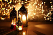 Ornamental Arabic lanterns with glittering bokeh lights. Burning candles on table glowing at night. Festive greeting card, invitation for Muslim holiday Ramadan Kareem. Golden Iftar background.