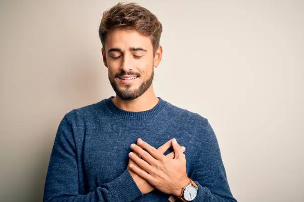 Young handsome man with beard wearing casual sweater standing over white background smiling with hands on chest with closed eyes and grateful gesture on face. Health concept.