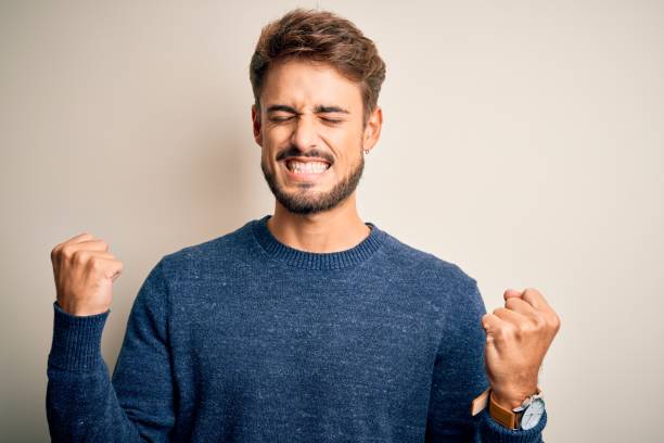 jeune homme beau avec la barbe utilisant le chandail occasionnel restant au-dessus du fond blanc très heureux et excité faisant le geste de gagnant avec les bras levés, souriant et criant pour le succès. concept de célébration. - men sweater excitement satisfaction photos et images de collection