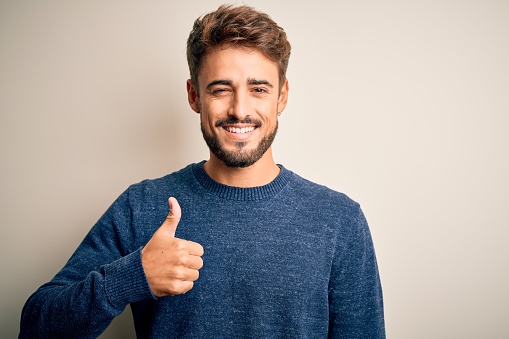Young handsome man with beard wearing casual sweater standing over white background doing happy thumbs up gesture with hand. Approving expression looking at the camera showing success.