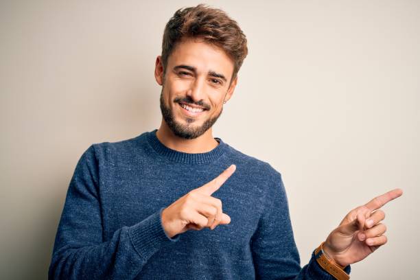 istock Young handsome man with beard wearing casual sweater standing over white background smiling and looking at the camera pointing with two hands and fingers to the side. 1212701566