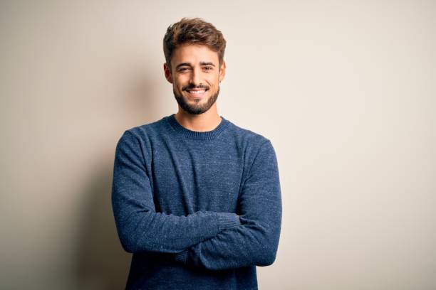白い背景の上に立っているカジュアルなセーターを着たひげを持つ若いハンサムな男は、カメラを見て腕を組んで微笑んでいます。ポジティブな人。 - スペイン 写真 ストックフォトと画像